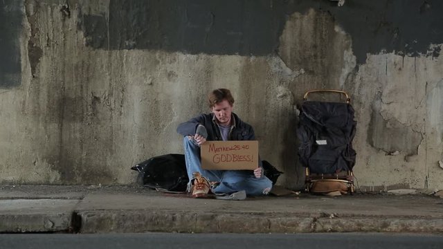 Wide Shot of a Homeless Man on the Sidewalk Under an Over Pass Holding a Sign with a Bible Verse and God Bless.