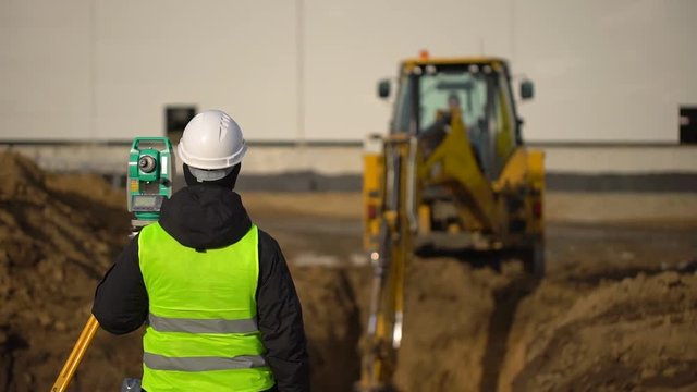 Male engineer, surveyor, builder, foreman, worker in uniform and hard hat working on construction site on background of working excavator machine. Engineer using tacheometer or theodolite for survey
