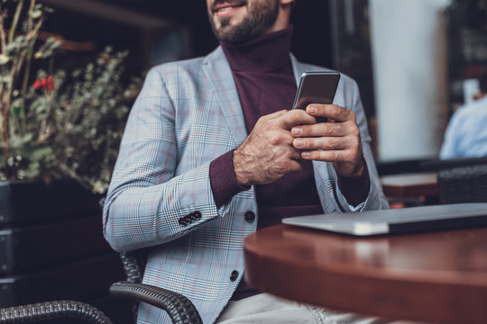 Cheerful Relaxed Young Man Sitting Outdoors At The Table With A Modern Convenient Device In His Hand And Smiling