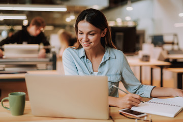 Fototapeta premium Portrait of beautiful smiling girl working with notebook and holding pencil while sitting at desk with cellphone and cup of coffee