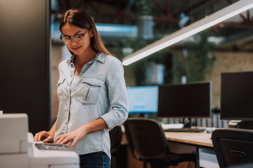Waist up portrait of charming woman standing near photocopier and smiling. Desk with computers on...