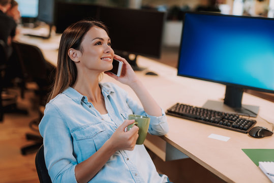 Side view portrait of charming woman holding cup of coffee and having phone conversation while sitting at office desk with computer. She is looking away and smiling
