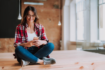 Portrait of charming girl holding smartphone while sitting on the floor. She is looking at phone display and smiling