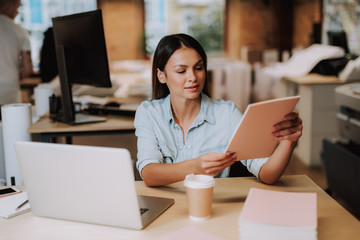 Fototapeta premium Portrait of charming woman sitting at table with laptop and holding working papers