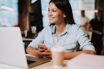 Portrait of beautiful woman with smartphone sitting at office desk with laptop. She is looking away and smiling