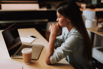 Lack of inspiration. Side view portrait of beautiful woman with cup of coffee sitting at office desk with laptop. She is keeping chin on hands and looking at display with serious expression