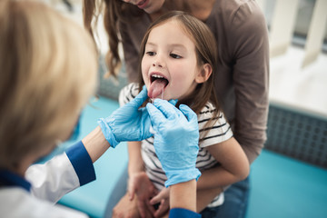 Concept of professional consultation and examination. Close up portrait of little girl with opening mouth being examined by pediatrician woman in medical office