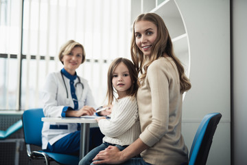 Concept of therapist consultation in healthcare system. Waist up low angle portrait of smiling little girl and her mother sitting in pediatrician cabinet