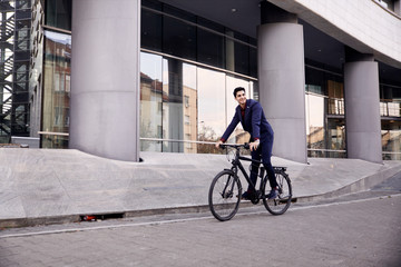one young man, 20-29 years old, wearing suit, looking smiling. riding, pedaling standing, fancy  bicycle. full length body. modern architecture building behind.