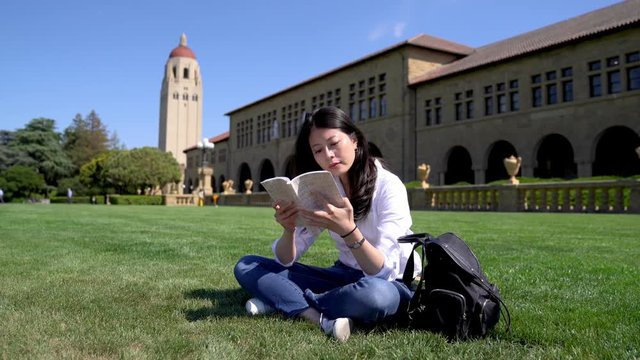 a diligent international student reading a book and sitting on the grass for a break during a full day of classes. 
