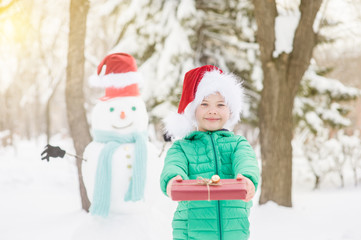 Fototapeta na wymiar smiling boy in red christmas hat holds gift box with snowman on background