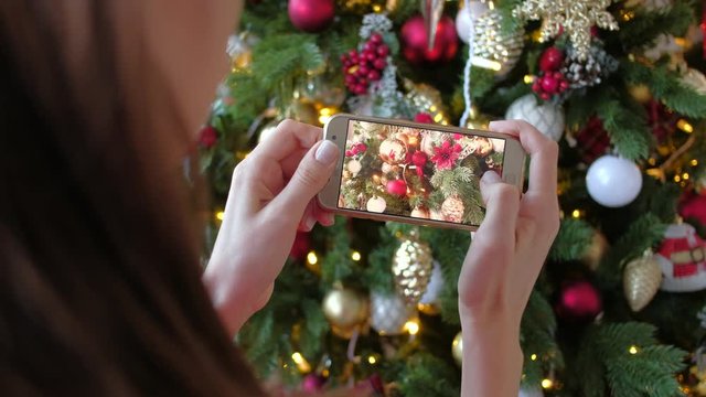 Beautiful woman photographing on smartphone christmas tree with toys at home. X-mas, winter holidays and people concept.
