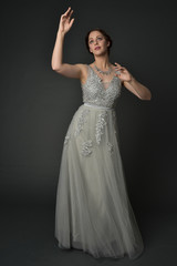 full length portrait of brunette  girl wearing long silver ball gown. standing pose on grey studio background.