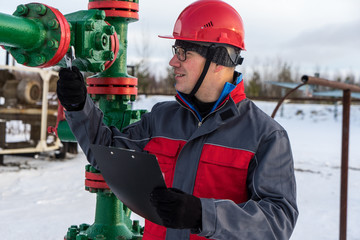 Man worker in the oilfield near pump jack and wellhead, wearing helmet and work clothes. Industrial site background. Oil and gas concept.