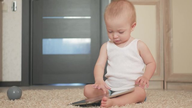 Baby boy with tablet computer sitting on the floor at home, indoors. Toddler handsome baby boy playing with screen device. Child plays with tablet technology at home.