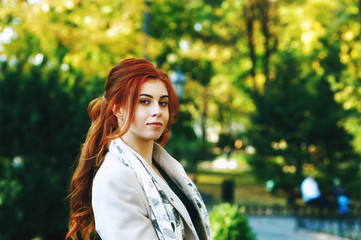 Portrait of a young beautiful fashionable red-haired woman with long curly hair , posing on the streets of the European city. The girl is dressed in a light coat