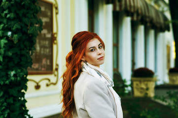 Portrait of a young beautiful fashionable red-haired woman with long curly hair , posing on the streets of the European city. The girl is dressed in a light coat