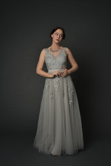 full length portrait of brunette  girl wearing long silver ball gown. standing pose on grey studio background.