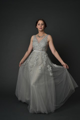 full length portrait of brunette  girl wearing long silver ball gown. standing pose on grey studio background.