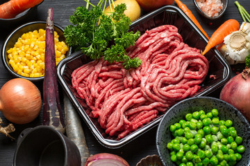 Raw minced meat in container with ingredients for shepherds pie with green peas, yellow corn, carrot, onion and seasonings, on black background, top view