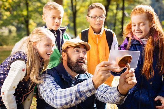 Smart teacher. Joyful bearded man taking photos of the mushroom while being surrounded by his pupils