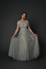 full length portrait of brunette  girl wearing long silver ball gown. standing pose on grey studio background.