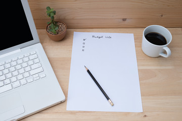 Paper check list for budget, laptop, coffee on wooden table.