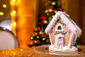 Gingerbread house in front of defocused lights of Christmas decorated fir tree. Holiday sweets. New Year and Christmas theme. Festive mood. Christmas card