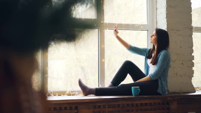 Cheerful Young Lady Is Making Online Video Call Sitting On Window Sill Looking At Smartphone Screen Smiling Waving Hand And Talking. Communication And Technology Concept.