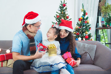 Asian family opening their presents together on Christmas morning. They are sitting on sofa in their living room in front of Christmas tree.
