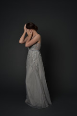 Fototapeta na wymiar full length portrait of brunette girl wearing long silver ball gown. standing pose with hands covering face on grey studio background.