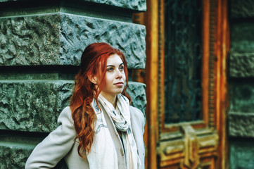 Fototapeta premium Portrait of a young beautiful fashionable red-haired woman with long curly hair , posing on the streets of the European city. The girl is dressed in a light coat