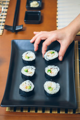 Hand of female chef placing japanese sushi rolls with rice, avocado and prawns on nori seaweed sheet over a black rectangular tray