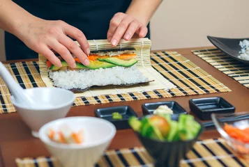 Foto op Aluminium Sushi bar Detail van handen van vrouwelijke chef-kok die Japanse sushi oprolt met rijst, avocado en garnalen op nori-zeewierblad