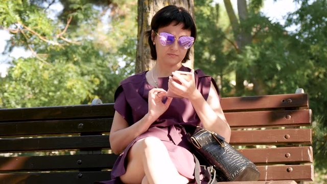 The girl sits on a park bench and uses a smartphone to communicate on social networks. Young brunette woman in sunglasses and dress.