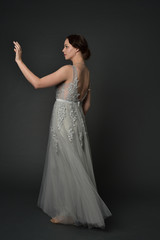 full length portrait of brunette  girl wearing long silver ball gown. standing pose on grey studio background.