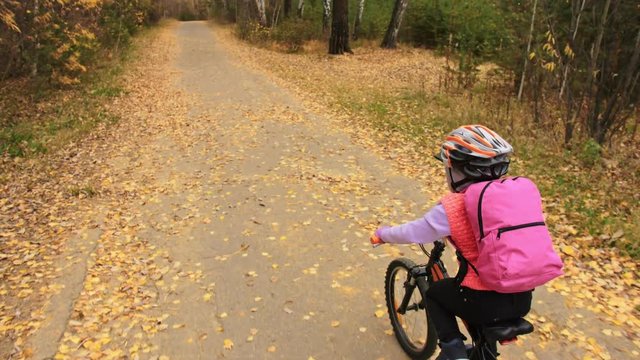 One caucasian children rides bike road in autumn park. Little girl riding black orange cycle in forest. Kid goes do bicycle sports. Biker motion ride with backpack and helmet. Mountain bike hardtail.