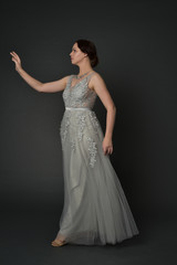 full length portrait of brunette  girl wearing long silver ball gown. standing pose on grey studio background.