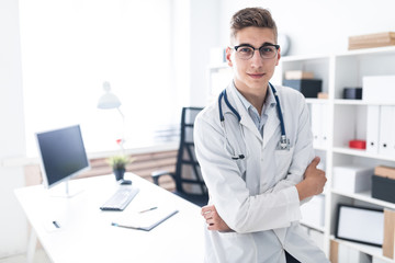 A young doctor in a white robe is leaning on the table in the office. He has a phonendoscope around his neck.