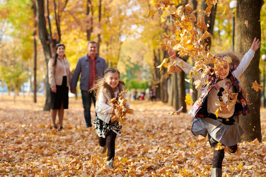 Happy Family Is In Autumn City Park. Children And Parents Running With Leaves.. They Posing, Smiling, Playing And Having Fun. Bright Yellow Trees.