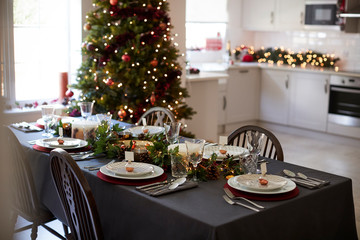 Christmas table setting with bauble name card holders arranged on plates and green and red decorations with Christmas tree and kitchen in the background