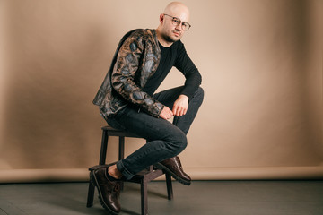 Lifestyle portrait of fashionable stylish bald man in glasses. Awesome man wearing biker leather jacket, black jeans, oxford shoes. Cool handsome well dressed boy on coffee paper studio background.