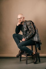 Lifestyle portrait of fashionable stylish bald man in glasses. Awesome man wearing biker leather jacket, black jeans, oxford shoes. Cool handsome well dressed boy on coffee paper studio background.