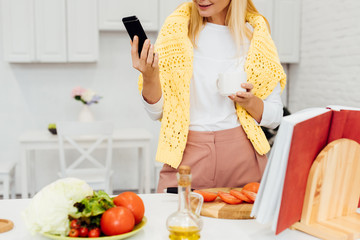 cropped view of blonde woman holding smartphone while cooking dinner