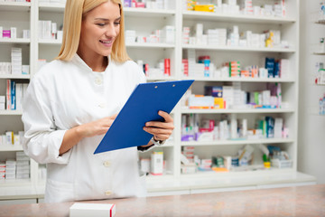 Happy young female pharmacist standing behind the counter with a clipboard, filling paperwork and writing. Medicine, pharmaceutics, health care and people concept