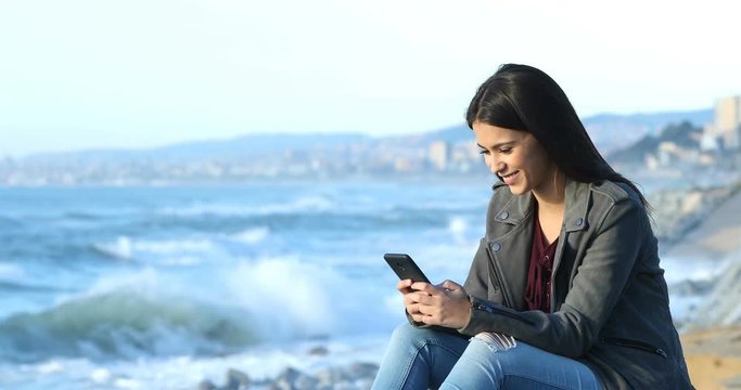 Happy teenage girl texting on smart phone sitting on the beach