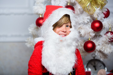 Boy kid dressed as santa with white artificial beard and red hat near christmas tree. Christmas santa claus costume for child. Christmas party with santa costume. Christmas tree ideas for kids