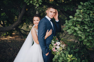 Happy newlyweds stand, embracing in a green garden with trees on the background of nature. Young bride and beautiful bride are cute smiling. Wedding photography. Stylish portrait.