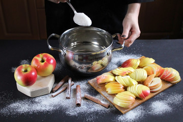Сhief cookes sugar syrup in a saucepan for apples baking. On black table lie ripe apples, powdered...