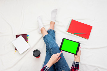 Girl in blue jeans and plaid shirt, is using tablet pc, women's feet, lying on white crumpled blanket. Women's hands, red manicure,  close up. Background with copy space, for advertisement. Top view